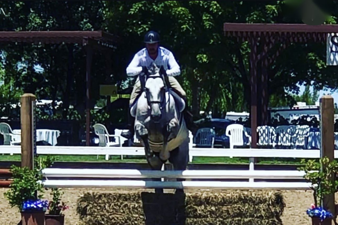 Ryan Oropeza riding Rendezvous over a hunter jump on a sunny day at a hay bale fence with flowers